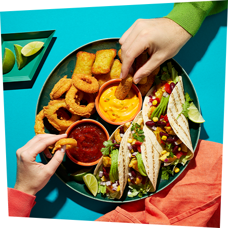 Ein Teller mit mexikanischen Tacos sowie Fingerfood und der Nacho Sauce und dem Paprika Relish von Develey als Dip